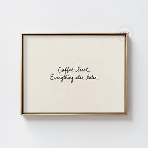 May include: A rectangular wall art print in a thin metallic frame featuring a cream-colored background with the black handwritten text Coffee first. Everything else later. centered in the middle of the frame with a small circular logo in the corner.