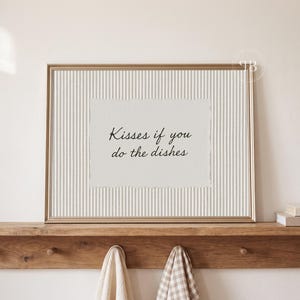 May include: A rectangular gold-toned frame containing a white paper insert with vertical pinstripes and the black text "Kisses if you do the dishes" printed in a cursive font, centered within the frame on a light-colored background.