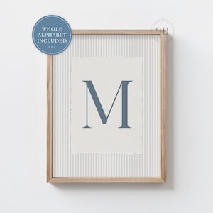 May include: A framed print displaying the letter "M" in a muted blue shade. The letter is centrally positioned on textured, off-white paper with a torn edge, set against a striped background. The frame is light wood, and a blue circle with text is in the upper left corner.