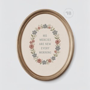 May include: An oval gold-toned frame containing a piece of fabric with an embroidered floral wreath in shades of blue and pink. The centre of the wreath features the embroidered text: HIS MERCIES ARE NEW EVERY MORNING in a dark grey font.