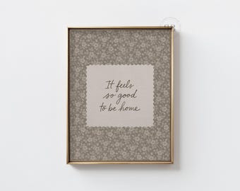 May include: A framed print with the text "It feels so good to be home." The artwork features a floral patterned background in shades of gray and a light beige center with the quote in elegant script. The frame is gold.