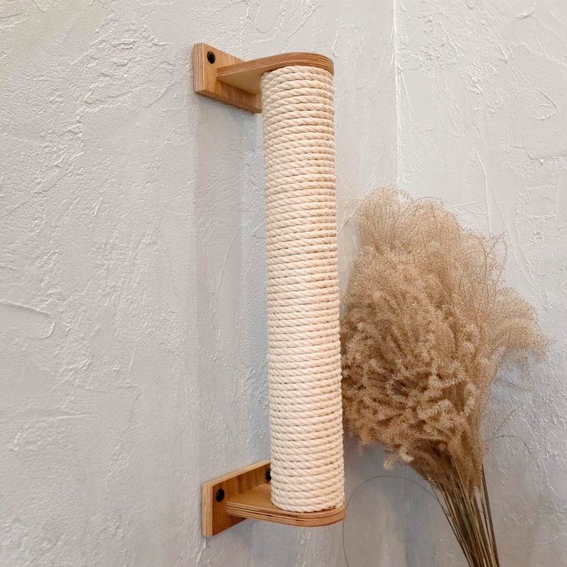May include: A wooden cat scratching post with a natural rope covering mounted on a wall. The post is in a corner and is attached to the wall with screws.