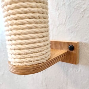 May include: A close-up of a wall-mounted cat scratching post made of natural wood and sisal rope. The post is attached to the wall with a wooden bracket and a black screw.