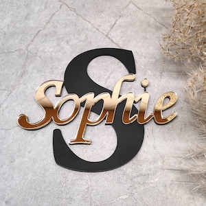 Può includere: Cartello con nome in acrilico a specchio dorato con il nome "Sophie" in un carattere corsivo. Il nome è ritagliato da uno sfondo in acrilico nero.