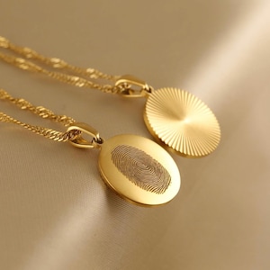 Peut inclure: Deux colliers pendentifs dorés. Un pendentif est de forme ovale avec un motif d'empreinte digitale, et l'autre est un pendentif rond avec un motif de rayons de soleil. Les deux pendentifs sont suspendus à de fines chaînes dorées.