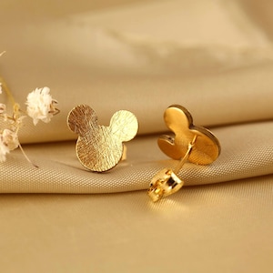 Può includere: Un paio di orecchini a lobo color oro a forma di silhouette di testa di topo. La superficie anteriore degli orecchini presenta una finitura spazzolata e testurizzata con sottili linee visibili, mentre il retro mostra il perno e una piccola chiusura a farfalla.