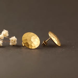 Op de afbeelding: Een paar goudkleurige oorstekers. Elke oorbel heeft een gehamerd, getextureerd oppervlak in een ovale vorm. De oorbellen worden gepresenteerd op een donkere achtergrond, met een takje witte bloemen in de linkerbovenhoek.