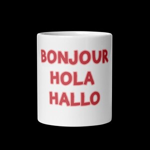 May include: White ceramic mug with the words "BONJOUR," "HOLA," and "HALLO" in red letters with a pink outline. The mug is cylindrical and stands against a black background.