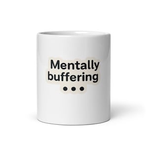 May include: A white ceramic mug with a black handle and the text "But First, Coffee" in a black, bold font. The mug is sitting on a wooden surface. The mug is approximately 4 inches tall.