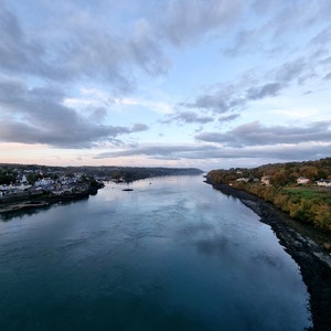 Puede incluir: Vista de un río con una ciudad a un lado y árboles al otro. El agua es de un azul profundo y el cielo es de un azul claro con nubes blancas.
