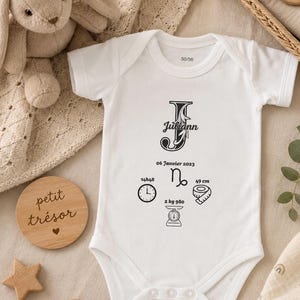 Peut inclure: Body bébé blanc avec le prénom "Juliann" dans une police décorative, ainsi que les détails de naissance. Le body comprend une horloge, un symbole du zodiaque, un mètre ruban (49 cm) et une balance (2 kg 980).