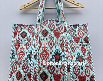 May include: A large tote bag with a colorful, patterned design. The bag features a light blue background with a repeating pattern of red, black, and cream-colored shapes. The handles and edges are trimmed with a contrasting light blue and white striped fabric.