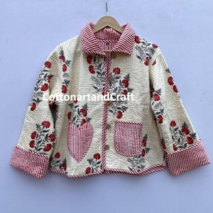 Peut inclure: Une veste matelassée crème avec une doublure rayée rouge et blanche. La veste est imprimée de fleurs rouges et de feuilles vertes. La veste a une poche en forme de cœur sur le devant et une fermeture à boutons.