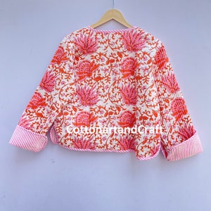 May include: A quilted jacket with a floral pattern in shades of pink and orange on a white background. The cuffs and trim feature a pink and white striped design. The jacket is hanging on a wooden hanger.