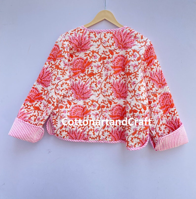 May include: A quilted jacket with a floral pattern in shades of pink and orange on a white background. The cuffs and trim feature a pink and white striped design. The jacket is hanging on a wooden hanger.