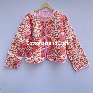 May include: A reversible quilted jacket with a floral pattern in shades of orange and pink on an off-white background. The jacket has a round neckline, long sleeves, and two pockets. The trim is pink and the lining is striped. The text "cottonartandcraft" is visible.