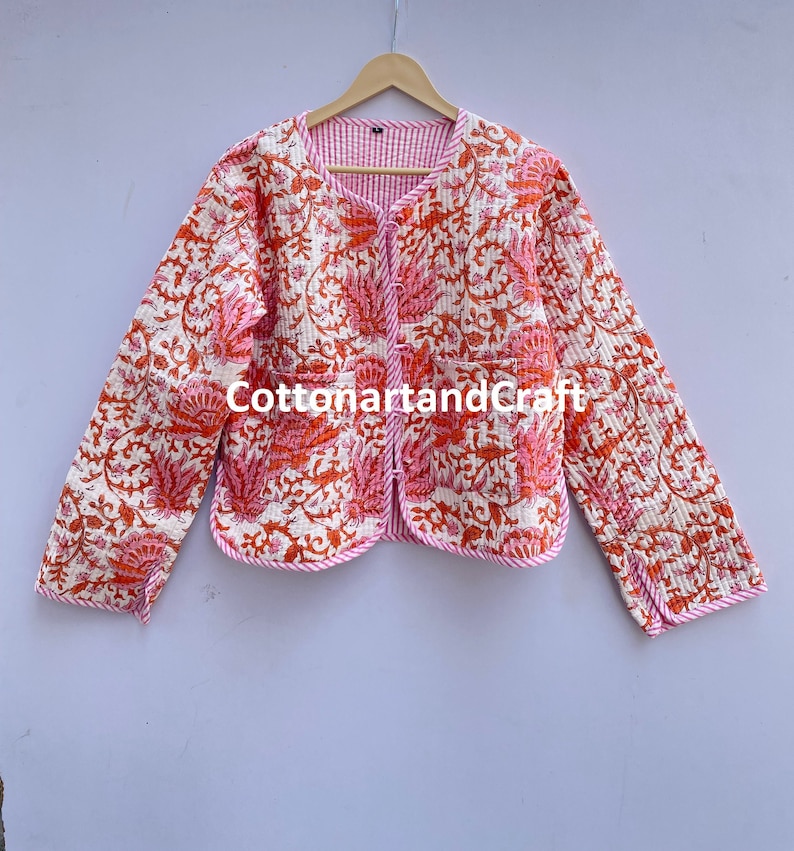 May include: A reversible quilted jacket with a floral pattern in shades of orange and pink on an off-white background. The jacket has a round neckline, long sleeves, and two pockets. The trim is pink and the lining is striped. The text "cottonartandcraft" is visible.