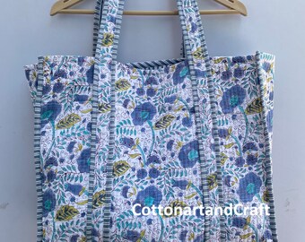 May include: A large white tote bag with blue and green floral print. The bag has gray and white striped handles and a gray and white striped trim. The bag is hanging on a wooden hanger. The text "CottonartandCraft" is visible on the bag.