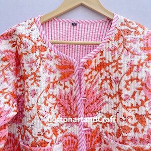 May include: A quilted jacket with a floral pattern in shades of pink and orange on a white background. The jacket has a striped pink lining and trim, with a button closure and two pockets. The text "cottonartandcraft" is visible.