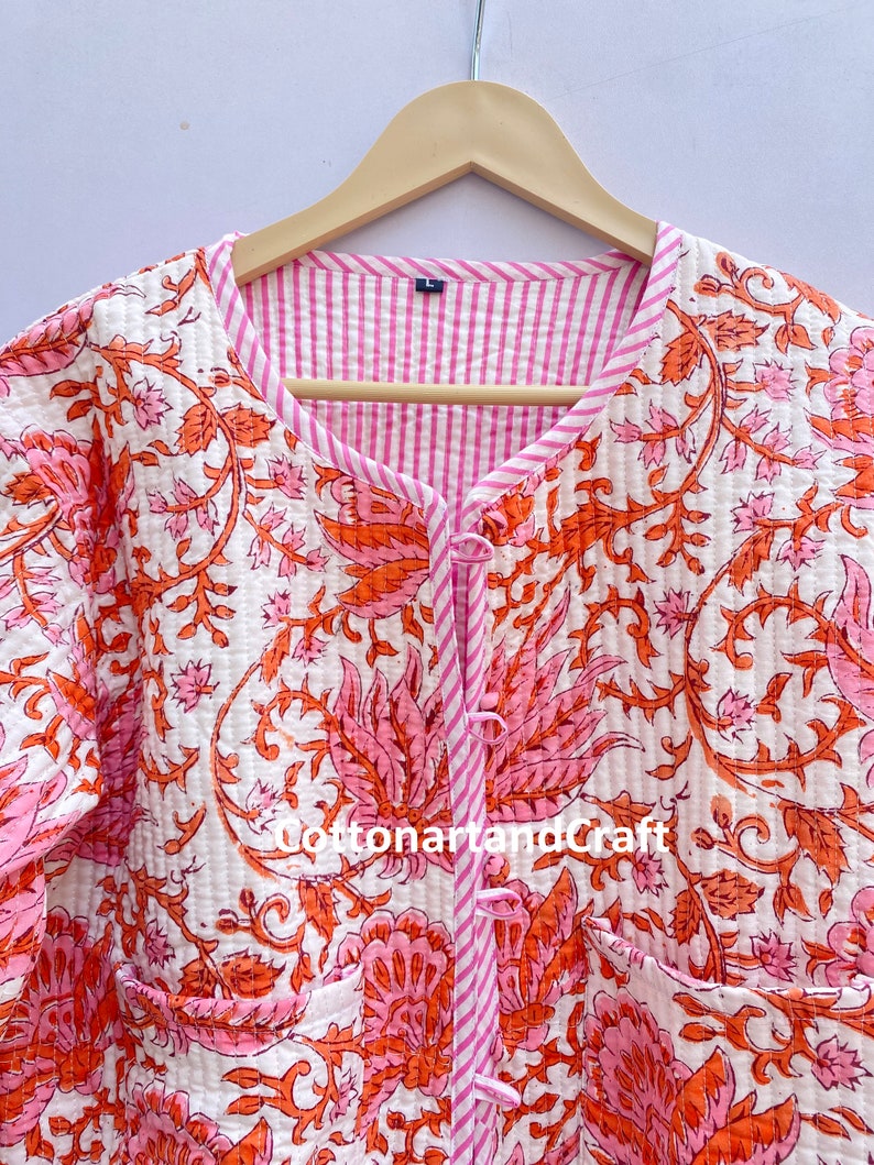 May include: A quilted jacket with a floral pattern in shades of pink and orange on a white background. The jacket has a striped pink lining and trim, with a button closure and two pockets. The text "cottonartandcraft" is visible.