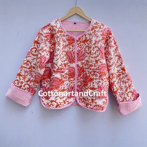 May include: A quilted jacket with a floral pattern in shades of pink and orange on a white background. The jacket has a round neckline, long sleeves, and a striped pink and white lining. The text "cottonartandcraft" is visible.