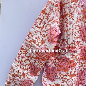 May include: A quilted jacket with a floral pattern in shades of pink and orange on a white background. The jacket has a pink and white striped trim and the text "cottonartandcraft".