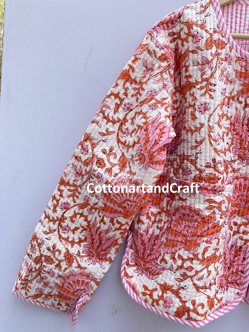 May include: A quilted jacket with a floral pattern in shades of pink and orange on a white background. The jacket has a pink and white striped trim and the text "cottonartandcraft".