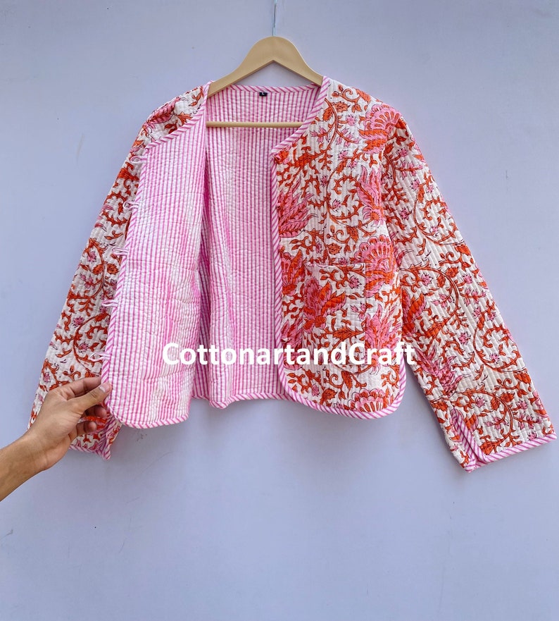 May include: A quilted jacket with a floral pattern in shades of orange and pink on a cream background. The jacket's interior is striped pink and white. The jacket is hanging on a wooden hanger.