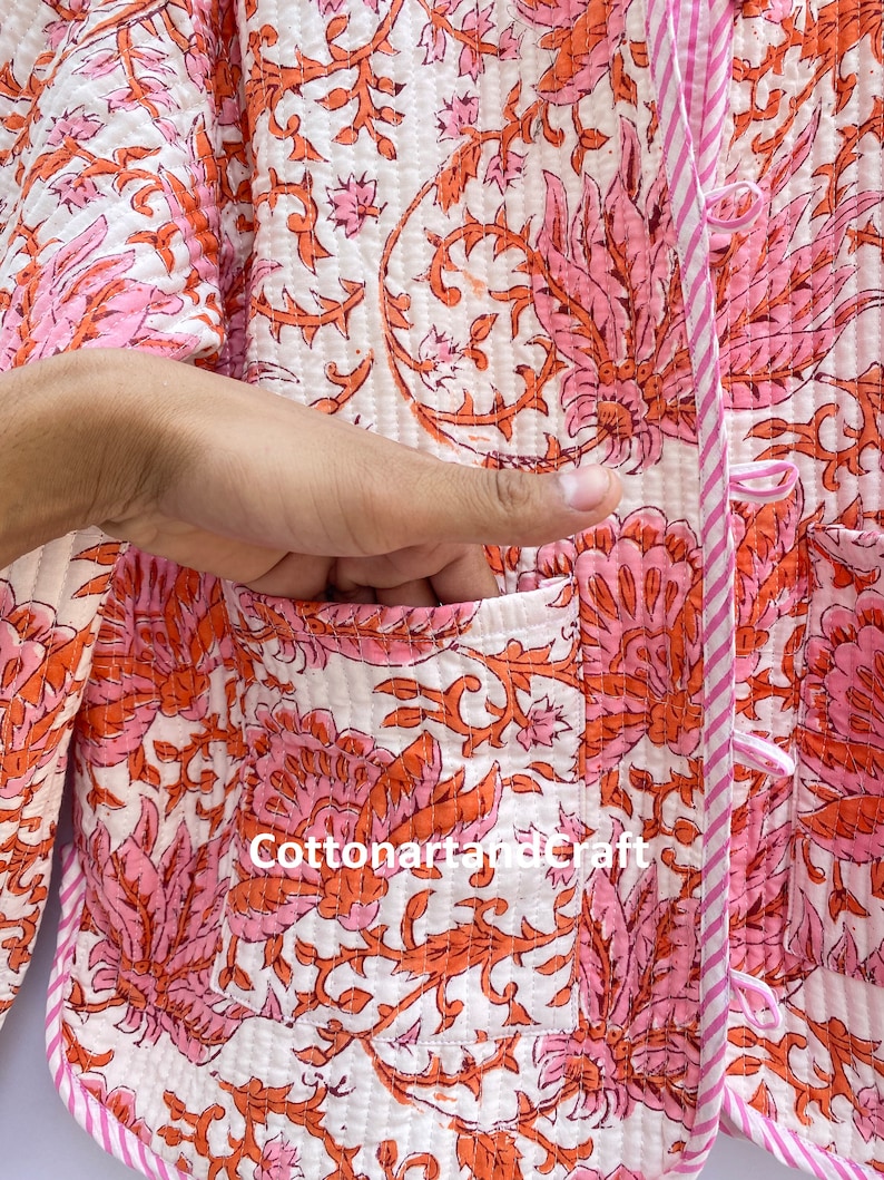 May include: A quilted jacket with a floral pattern in shades of pink and orange on a white background. The jacket has a pocket and is trimmed with pink and white striped fabric. The text "Cottonartandcraft" is visible on the jacket.