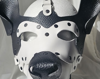 May include: A black and white leather pup hood with a textured snout, eye holes, and ear-like flaps. The hood has a strap with a buckle and is designed to fit over the head.