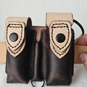 May include: A handmade leather pouch with two compartments. The pouch is dark brown with lighter tan accents on the flaps, secured by black buttons. A black metal ring is attached at the bottom. The stitching is visible.