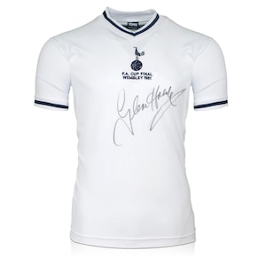 Könnte beinhalten: Weißes Fußballtrikot mit dunkelblauem Kragen und Besatz. Das Trikot trägt das Wappen von Tottenham Hotspur und den Text "F.A. Cup Final Wembley 1981". Das Trikot ist von Glenn Hoddle signiert.