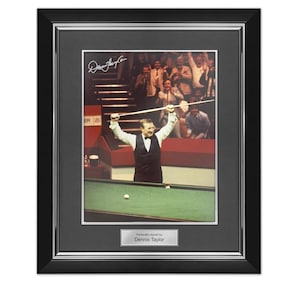 May include: Framed photograph of a man celebrating a snooker victory, holding a cue stick above his head. The image is signed by Dennis Taylor and set in a black frame with a silver border. The photograph shows a green snooker table and a crowd of people in the background.