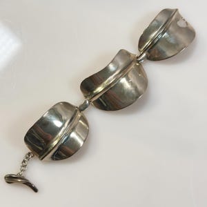 May include: A silver-toned metal bracelet featuring three large, curved, leaf-shaped links connected by small circular rings. The bracelet includes a short chain segment and a T-bar toggle clasp at one end, with a polished, reflective surface.