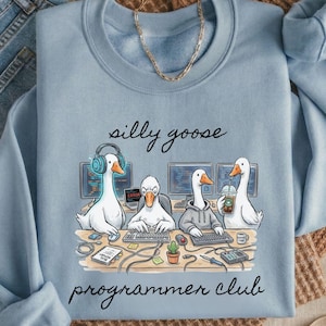 May include: A pale blue sweatshirt with a cartoon design of geese working on computers. The words "silly goose programmer club" are written above and below the image. The sweatshirt is shown with blue jeans and a gold necklace.