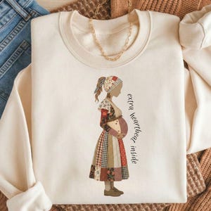 May include: Cream-colored sweatshirt with a graphic of a pregnant woman in a patchwork dress. The text "extra heartbeat inside" is printed vertically on the right side. The sweatshirt is laid on a brown sweater and next to blue jeans.