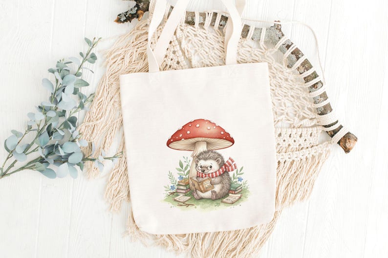 May include: A cream-colored tote bag featuring a whimsical illustration of a hedgehog reading under a red-capped mushroom. The hedgehog wears a scarf and glasses, surrounded by books and greenery. The bag has sturdy handles.