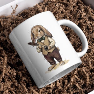 May include: White ceramic mug featuring a watercolor illustration of a rabbit dressed in a vest, bow tie, and pants, holding a quill and inkwell. The mug is set in a white box with brown paper filler.