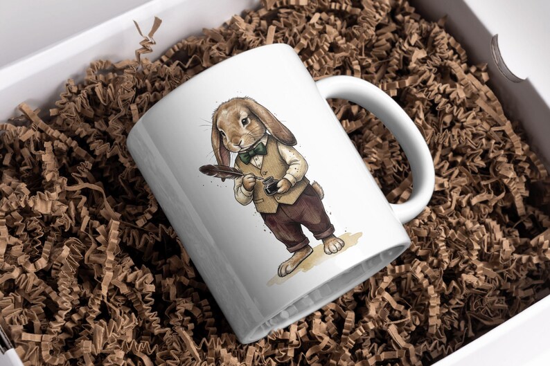 May include: White ceramic mug featuring a watercolor illustration of a rabbit dressed in a vest, bow tie, and pants, holding a quill and inkwell. The mug is set in a white box with brown paper filler.