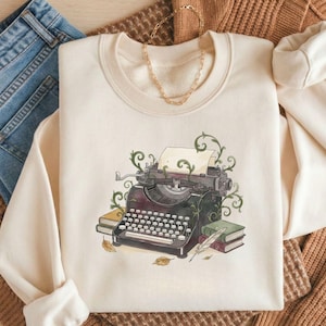 May include: Cream-colored sweatshirt featuring a vintage typewriter illustration with green vines, a sheet of paper, and books. The design includes a quill pen and autumn leaves. The sweatshirt is paired with blue jeans and a brown sweater.