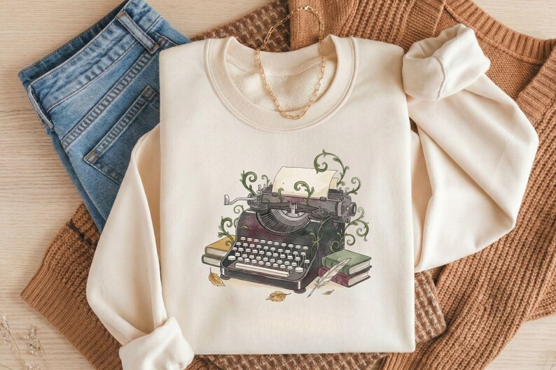May include: Cream-colored sweatshirt featuring a vintage typewriter illustration with green vines, a sheet of paper, and books. The design includes a quill pen and autumn leaves. The sweatshirt is paired with blue jeans and a brown sweater.