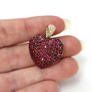 May include: A heart-shaped pendant featuring a dense pavé setting of numerous small, deep red circular gemstones. The pendant includes a triangular bail at the top, which is encrusted with tiny, clear white sparkling stones set in gold.