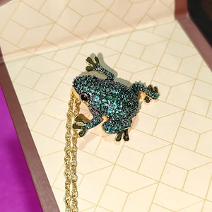May include: A gold-toned necklace featuring a frog pendant. The frog is covered in emerald-green stones with black eyes. The necklace chain is a delicate gold color. The pendant is a detailed, jewel-toned accessory.