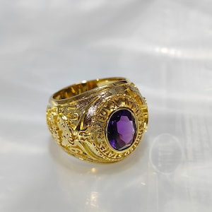 May include: A gold ring featuring a central oval, deep purple gemstone. The ring's band is adorned with detailed engravings and text. The ring is displayed against a white backdrop, highlighting its design and craftsmanship.
