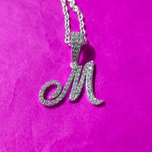 May include: A silver necklace featuring a pendant shaped like the letter "M", embellished with small, clear gemstones. The chain and pendant are set against a vibrant pink backdrop.