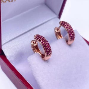 Peut inclure: Une paire de créoles en or rose serties de petites pierres roses éclatantes. Les boucles d'oreilles sont présentées dans un écrin à bijoux doublé de velours blanc avec un extérieur rouge profond, mettant en valeur le métal et les pierres.