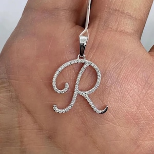 Könnte beinhalten: Ein silberner Anhänger in Form des Buchstabens "R", besetzt mit kleinen, funkelnden Edelsteinen. Der Anhänger hängt an einer dünnen Silberkette und bildet ein zartes und elegantes Schmuckstück.