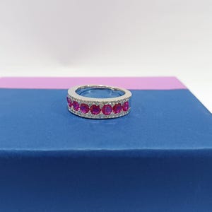 Peut inclure: Une bague en argent ornée d'une rangée de pierres précieuses rouges vives, entourée de petites pierres claires. La bague est présentée sur une surface bleue, mettant en valeur son design élégant et ses détails scintillants. Un bijou.