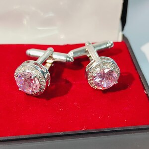 May include: Silver cufflinks featuring large, round, pink gemstones. Each cufflink has a halo of smaller, clear stones. Presented in a red velvet-lined box. The cufflinks are a classic accessory.
