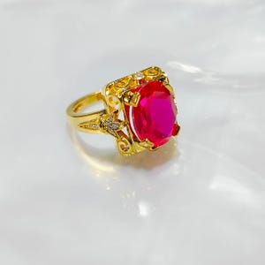 Puede incluir: Un anillo dorado con una gran piedra preciosa rosa brillante de talla ovalada. El anillo tiene un diseño de filigrana ornamentado alrededor de la piedra preciosa y a lo largo de la banda, con pequeñas piedras de acento. El anillo está sobre un fondo blanco.